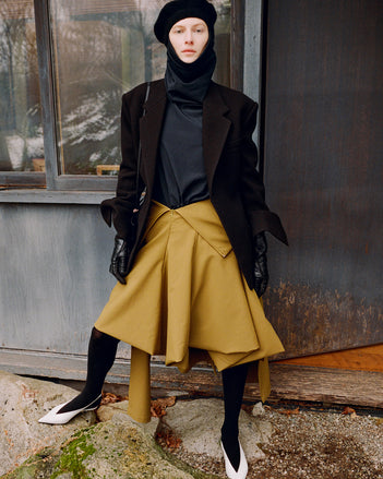 Model standing against the side of a window wearing Daria Jacket in Black Wool Scuba, Solene Top in Anthracite Silk Viscose, Tiana Dress in Fatigue Technical Canvas, Beret in Black Wool Knit, Reade Shoulder Bag in Black Paper Calf, and Slash Slingbacks in Optic White Spazzolato