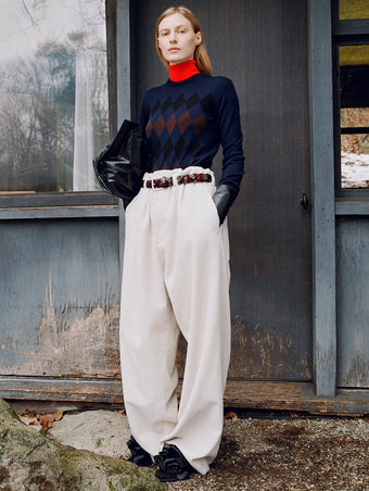 Model standing against the side of a rustic house wearing Vivian Sweater in Navy Argyle, Fox Sweater in Red Viscose, Salome Pant in Ecru Corduroy, Days Carryall in Black Paper Calf, Braid Belt in Bordeaux Croc Embossed Calf, and Tee Souvenir Sandals in Black Satin