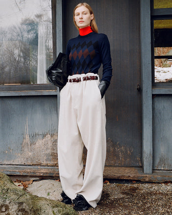 Model standing against the side of a rustic house wearing Vivian Sweater in Navy Argyle, Fox Sweater in Red Viscose, Salome Pant in Ecru Corduroy, Days Carryall in Black Paper Calf, Braid Belt in Bordeaux Croc Embossed Calf, and Tee Souvenir Sandals in Black Satin