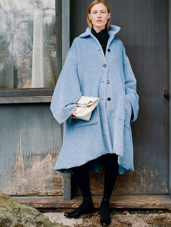 Model standing against the side of a rustic house wearing Verretta Coat in Sky Blue Alpaca Coating, Ysabelle Top in Anthracite Silk Viscose, Reade Shoulder Bag in Optic White Paper Calf, and Uma Tie Pumps in Black Slick Calf