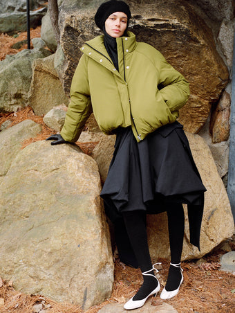Model leaning against stone boulder wearing Laney Jacket in Moss Stretch Nylon, Tiana Dress in Black Technical Canvas, Solene Top in Anthracite Silk Viscose, Beret in Black Wool Knit, and Uma Tie Pumps in Optic White Slick Calf