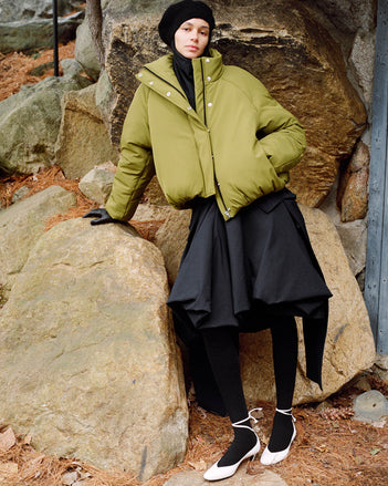 Model leaning against stone boulder wearing Laney Jacket in Moss Stretch Nylon, Tiana Dress in Black Technical Canvas, Solene Top in Anthracite Silk Viscose, Beret in Black Wool Knit, and Uma Tie Pumps in Optic White Slick Calf
