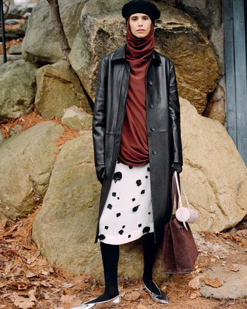 Model standing against stone boulders wearing Nissi Coat in Black Semigloss Leather, Solene Top in Mahogany Silk Viscose, Lex Skirt in White/Black Embroidered Viscose Georgette, Silo Bag in Light Grey Croc Embossed Calf, Days Tote in Chocolate Suede, and Slash Flats in Silver Specchio