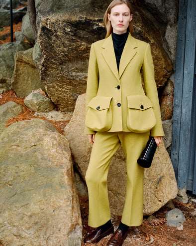Model standing against stone wall wearing Silva Jacket in Fatigue Wool Scuba, Ismene Pant in Fatigue Wool Scuba, Ysabelle Top in Anthracite Silk Viscose, Silo Clutch in Black Shiny Calf, and Albers Loafer in Cedar Shiny Calf