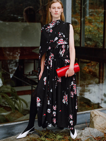 Model standing against glass windows wearing Ysabelle Dress in Black Printed Silk Viscose, Silo Clutch in Red Shiny Calf, and Slash Slingbacks in Optic White Spazzolato