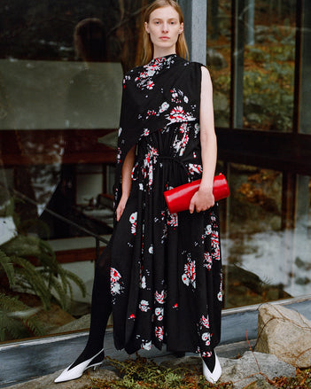 Model standing against glass windows wearing Ysabelle Dress in Black Printed Silk Viscose, Silo Clutch in Red Shiny Calf, and Slash Slingbacks in Optic White Spazzolato