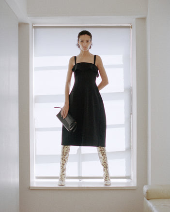 Model standing in a white windowsill wearing a black dress and holding the Tate Clutch