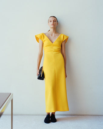 Model wearing the Mariam dress leaning against white wall holding the Armory Clutch