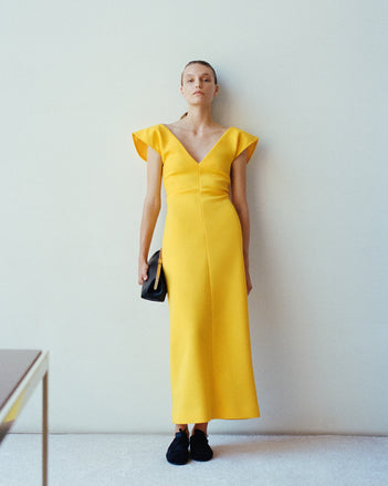 Model wearing the Mariam dress leaning against white wall holding the Armory Clutch