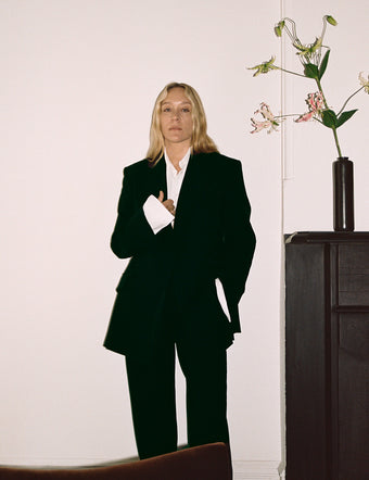 Image of Chloe Sevigny wearing black Wool Stretch Suiting Jacket and Trousers leaning against wall against dresses and flowers