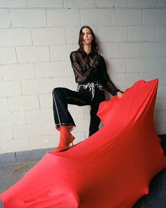 Model Jeanne Cadieu wearing the Smithson top in Black Organza Knit with the black multi Ryman jean from FW24, leaning against a beige brick wall and propping her leg on a motorcycle covered with a red motorcycle covering
