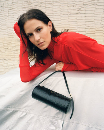 Cropped image of model laying across covered car wearing Paula Dress in Sheer Crepe Jersey in red, carrying Silo Bag in Ostrich Embossed Calf in black