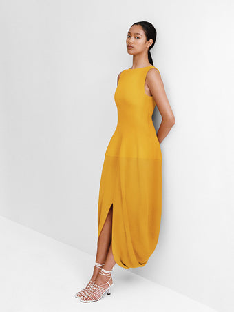 Editorial Image of model leaning against a white wall wearing the Sukki Dress in Draped Viscose in Marigold with the Tee Knot Sandals in Nappa in White