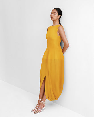 Editorial Image of model leaning against a white wall wearing the Sukki Dress in Draped Viscose in Marigold with the Tee Knot Sandals in Nappa in White