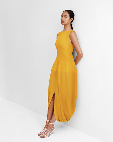 Editorial Image of model leaning against a white wall wearing the Sukki Dress in Draped Viscose in Marigold with the Tee Knot Sandals in Nappa in White