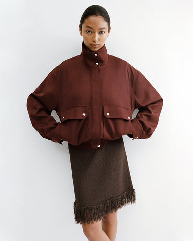 Proenza Schouler Editorial image of model standing against white wall wearing the Aviva Jacket in Wool Gabardine in Oxblood with the Saski Dress in Cotton in Textured Fringe in Earth