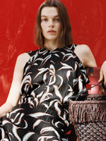Proenza Schouler campaign image of model sitting in front of red background wearing Sybella Dress in Metalasse Silk Linen in Black Multi with the Bucket Bag in Raffia in plum