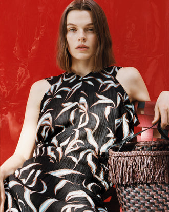 Proenza Schouler campaign image of model sitting in front of red background wearing Sybella Dress in Metalasse Silk Linen in Black Multi with the Bucket Bag in Raffia in plum