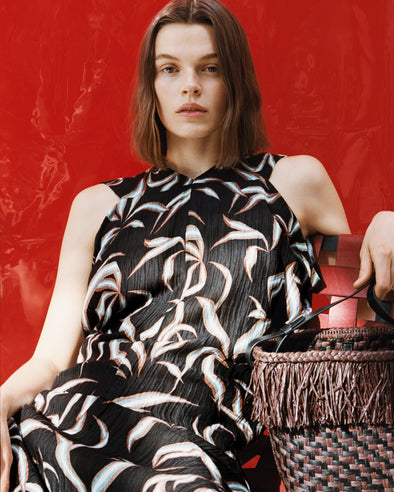 Proenza Schouler campaign image of model sitting in front of red background wearing Sybella Dress in Metalasse Silk Linen in Black Multi with the Bucket Bag in Raffia in plum