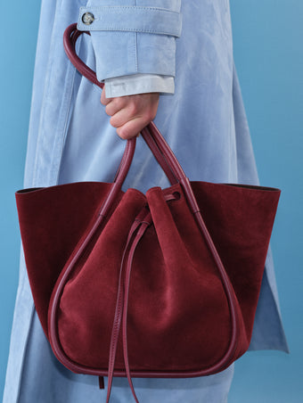 Image of model wearing Ramona Coat in Suede in light blue holding Large Ruched Tote in Soft Suede in Merlot with blue background. 