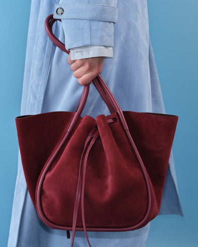 Image of model wearing Ramona Coat in Suede in light blue holding Large Ruched Tote in Soft Suede in Merlot with blue background. 
