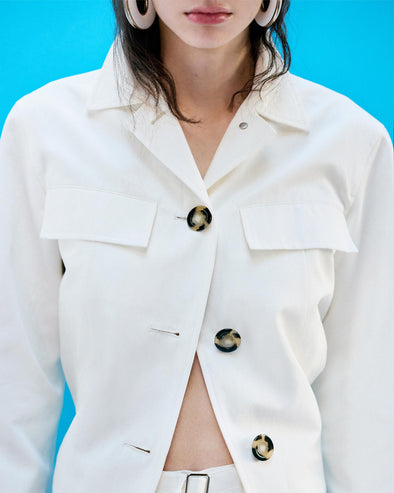 Cropped image of model standing in front of a blue backdrop, wearing Thelma Jacket in Cotton Linen Suiting in White
