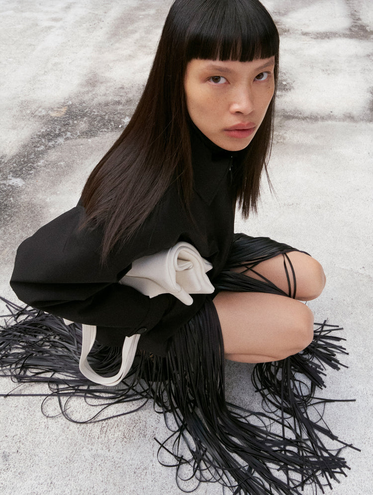 Image of model crouching on concrete ground wearing Sibyl Jacket in Stretch Nylon in black with Veda Skirt in Faux Leather Fringe in black