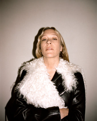 Image of Chloe Sevigny wearing a black leather jacket with a white fur collar against an off-white background, arms folded across her chest