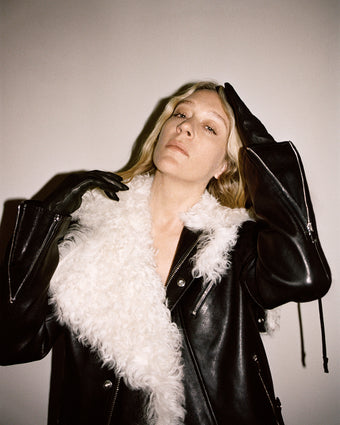 4x5 image of Chloe Sevigny standing against an off white background wearing a black leather jacket with a white fur collar, her left arm raised touching the top of her head