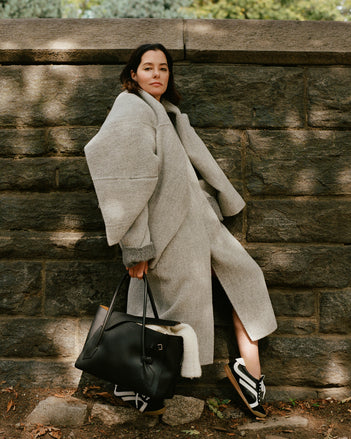 Image of Parker Posey against brick wall, wearing Albers Coat in Textured Double Face Wool in light grey multi and Track Sneakers in black/white, carrying Tate Bag in black