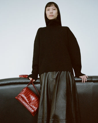 Cropped image of a model wearing Sandra Turtleneck in Eco Doubleface Cashmere in black, Moore Skirt in Glossy Leather in black, carrying a City Bag in Eel in rosewood.
