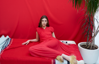 Model laying against tonal red backdrop, wearing Rumi Dress in Pleated Jersey in scarlet and Tee Thong Feather Sandals in resin