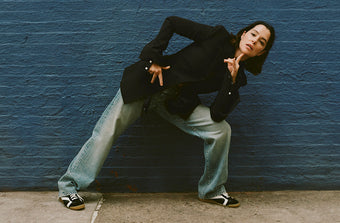 Image of Parker Posey against blue brick wall, wearing Lark Blazer in Wool Gabardine Suiting in black, Ellsworth Jean in classic wash, and Track Sneakers in black/white