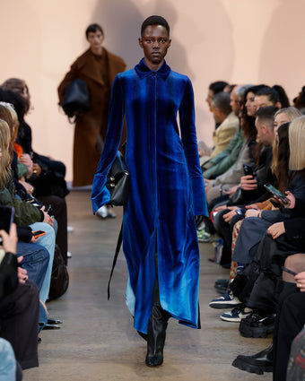 4x5 image of model Alaato Jazyper walking a runway wearing the blue velvet ice dye shirt dress with black cone boots and a black bar bag, with spectators legs on the right and left sides of the image and another model in the distant background