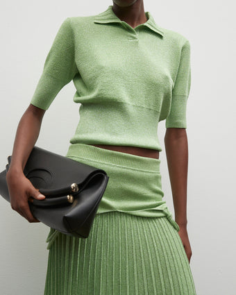 4x5 cropped image of a model wearing green metallic lurex knit polo top and dress with the top folded over the skirt, holding a black bar bag in her right hand against her right hip, standing against a white background