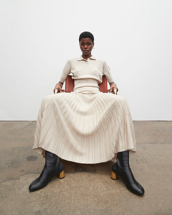 4x5 image of a model sitting down in a rust colored chair against a white background and brown linoleum floor, the model is wearing the champagne lurex knit polo top and champagne metallic knit dress as well as black sculpted boots with a gold heel