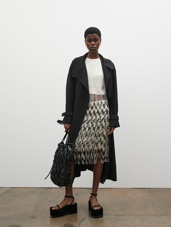 PF23 Collection Look 18: Black Marocain Trench Coat, Ecru / White Graphic Embroidered Fringe Top, Black / Ecru Graphic Embroidered Fringe Skirt, Black Macrame Drawstring Tote, and Black Velvet Square Platform Sandals