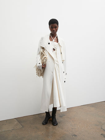 PF23 Collection Look 25: White Marocain Trench Coat, White Glossy Leather Dress, Ecru Macrame Drawstring Tote, and Black Sculpt Ankle Boots with Gold Heel