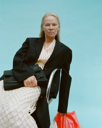 Image of Pamela Anderson sitting on metal chair in Tauba Dress in Satin Ribbon in resin and Devon Jacket in Viscose Wool in black, holding black Nylon City Messenger Bag