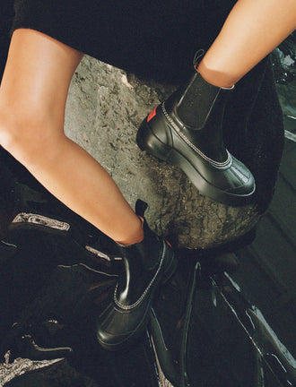 Image of model sitting on a rock wearing SOREL x Proenza Schouler Caribou Chelsea Boots in black/chalk