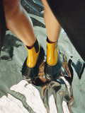 Aerial image of model wearing SOREL x Proenza Schouler Caribou Chelsea Boots in ochre/black, standing on silver floor