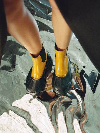 Aerial image of model wearing SOREL x Proenza Schouler Caribou Chelsea Boots in ochre/black, standing on silver floor