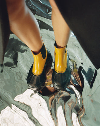 Aerial image of model wearing SOREL x Proenza Schouler Caribou Chelsea Boots in ochre/black, standing on silver floor
