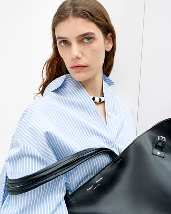 Cropped image of model wearing Isla Dress in Stripe Cotton Silk Poplin in pale blue multi and holding Days Carryall in Smooth Calf in black 