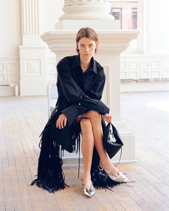 Model sitting on white chair, wearing Addy Shirt in Compact Cotton Poplin and Phia Skirt in Leather Silk Fringe in black with Slash Crossover Slingback Pumps in silver, carrying Tetra Bag in black