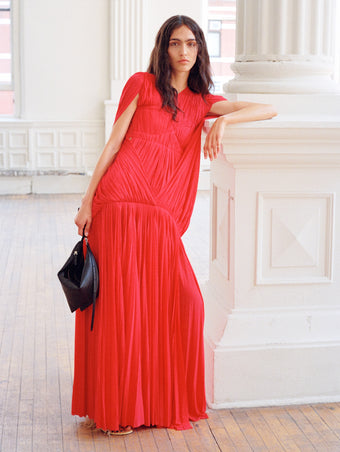 Model leaning against white column wearing Rumi Dress in Pleated Jersey in scarlet, carrying Tetra Shoulder Bag in Nappa in black