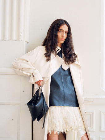 Model wearing Sabina Coat in Lacquered Leather in ecru and Noa Top in Textured Leather in black, carrying Tetra Bag in black