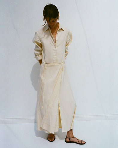 Model looking down at her feet with her arms tucked behind her, wearing Lato Wrap Dress in Linen Slub in light wheat