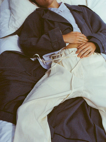 Cropped image of model laying across bed with hands folded on her stomach, wearing Sakia Coat in Wool Melange in Denim with Magnus Pant in Linen Slub in light wheat