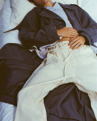 Cropped image of model laying across bed with hands folded on her stomach, wearing Sakia Coat in Wool Melange in Denim with Magnus Pant in Linen Slub in light wheat